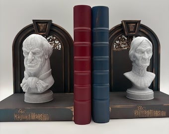 May include: Bookends featuring a pair of red and blue books flanked by busts of a stern-faced man. The bookends have a dark brown arched design with the text "The Haunted Mansion" on the base.