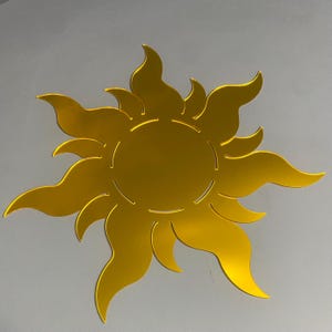 May include: A golden yellow sun-shaped decorative item with stylized flame-like rays. The sun has a circular center and is set against a light gray background. The sun's rays are curved and pointed.