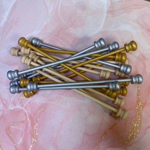 May include: A bundle of wooden dowels with gold and silver painted tips. The dowels are arranged in a criss-cross pattern.