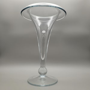 May include: A clear glass vase with a flared top and a long, slender stem. The vase has a wide, circular base and a bulbous section near the bottom of the stem. The top of the vase has a rounded, flared shape, and the glass is smooth and transparent.