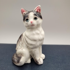 May include: A ceramic cat figurine in a seated position. The cat is white with grey and black markings, blue eyes, and pink details on the ears and nose. The figurine is detailed and has a glossy finish.
