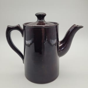 May include: A dark brown ceramic coffee pot with a lid and a curved handle. The spout is long and angled. The pot has a glossy finish and is set against a white background. The coffee pot is a vintage style.
