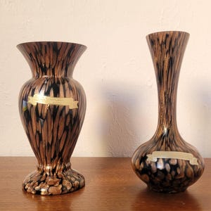 May include: Two glass vases featuring a black and metallic copper speckled pattern. The left vase has a wide, flared rim and a bulbous body, while the right vase has a slender neck and a rounded base. Both vases display a gold-coloured label.