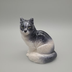 May include: A ceramic cat ornament in a seated pose. The cat is predominantly grey and white with yellow eyes. The figurine has a glossy finish, possibly porcelain. The cat is sitting upright, with its front paws together, and appears to be looking forward.