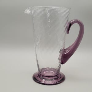 May include: A clear glass jug with a purple handle and base. The jug features a pouring spout and a decorative swirl pattern. The handle and base are a solid purple colour. The jug is suitable for serving drinks.