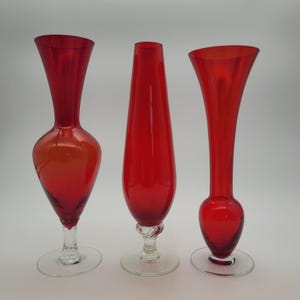 May include: Three red glass vases of different shapes. The vase on the left has a bulbous body and a flared top. The middle vase is tall and slender, while the one on the right has a rounded base and a flared top. All have clear glass bases.