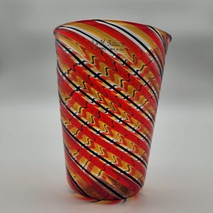 May include: A hand-blown glass tumbler with a striking, swirled pattern. The tumbler showcases alternating bands of red, gold, white, and black, forming a dynamic design. The glass is marked "J.M. Ballarin HAND MADE IN ITALY".