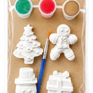 May include: A craft kit containing four white plaster figures shaped like a Christmas tree, a gingerbread person, a snowman, and a gift box. The set includes a blue paintbrush and three small paint pots in green, red, and brown, all packaged in a clear plastic bag on a brown cardboard backing.