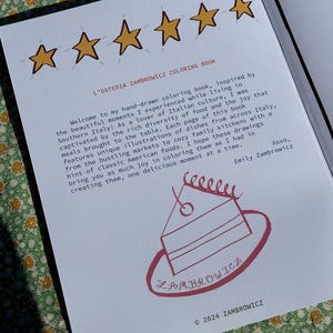 May include: Open coloring book page with text and illustrations. The title reads "L'Osteria Zambrowicz Coloring Book." The page includes a welcome message, a cake illustration, and the author's signature. The book is inspired by Italian culture and food.