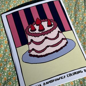 May include: A coloring book featuring a cartoon cake illustration with three strawberry toppings. The cake is on a blue plate, set against a pink and navy striped background. The book's title, "L'OSTERIA ZAMBROWICZ COLORING BOOK", is printed at the bottom.