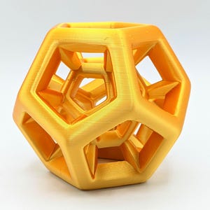 Puede incluir: Un objeto geométrico dorado impreso en 3D con un diseño complejo y abierto. El objeto tiene una forma multifacética con estructuras internas visibles y una superficie texturizada. El color es uniforme y el objeto está sobre un fondo blanco liso.