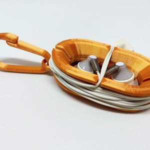 Puede incluir: Soporte para auriculares naranja impreso en 3D con un mosquetón. El soporte contiene auriculares blancos y un cable blanco enrollado. El soporte tiene forma ovalada con una ranura para sujetar los auriculares y el cable. El mosquetón está sujeto al soporte.