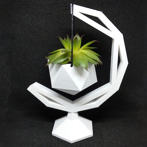 Op de afbeelding: Een witte geometrische plantenpot met een vetplant die aan een zwart koord hangt. De pot hangt aan een witte geometrische standaard.