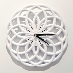 May include: White, geometric wall clock with black hands and a black and gold center. The clock has a decorative, openwork design. The clock is mounted on a white wall.