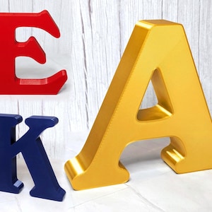 May include: Three dimensional block letters including a red letter E, a dark blue letter K, and a large gold letter A. Each letter features a serif font style with a smooth finish and stands upright on a flat surface, displaying depth and thickness.
