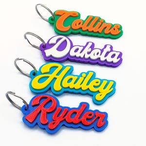 May include: Four colorful name keychains with silver key rings. The names are "Collins" in orange and green, "Dakota" in white and purple, "Hailey" in yellow and blue, and "Ryder" in red and blue. The keychains are layered with a 3D effect.