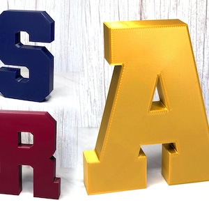 May include: Three large, block-style letters: a navy blue "S", a maroon "R", and a gold "A". Each letter is freestanding and has a geometric design. The letters are likely decorative or for display.