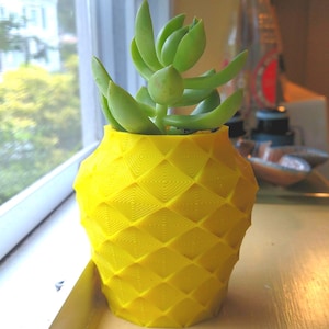 May include: A yellow pineapple-shaped plant pot with a geometric design, containing a green succulent. The pot is made of a solid material and is placed on a wooden surface near a window.