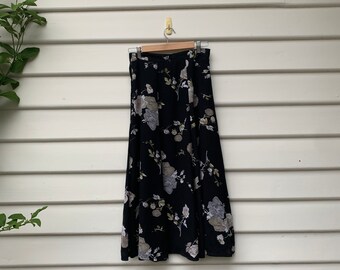 Könnte beinhalten: Ein schwarzer Midirock mit einem Blumenmuster aus gedeckten grauen, beigen und weißen Blüten sowie grünen Blättern. Der Rock hat einen geraden Bund, hängt an einem Holzkleiderbügel und zeigt eine fließende Silhouette mit leichtem A-Schnitt.