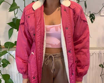 May include: A long-sleeved, magenta-colored jacket featuring a white, fuzzy, sherpa-style lining. The jacket has a front zipper closure, metal snap buttons, and a high collar. It is worn over a light pink crop top and tan drawstring sweatpants.