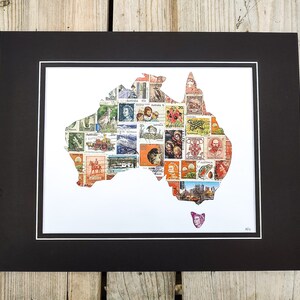 May include: A framed artwork featuring a map of Australia composed of vintage postage stamps. The stamps are of various colors and designs, set against a white background, and matted in black. The artwork is displayed on a wooden surface.