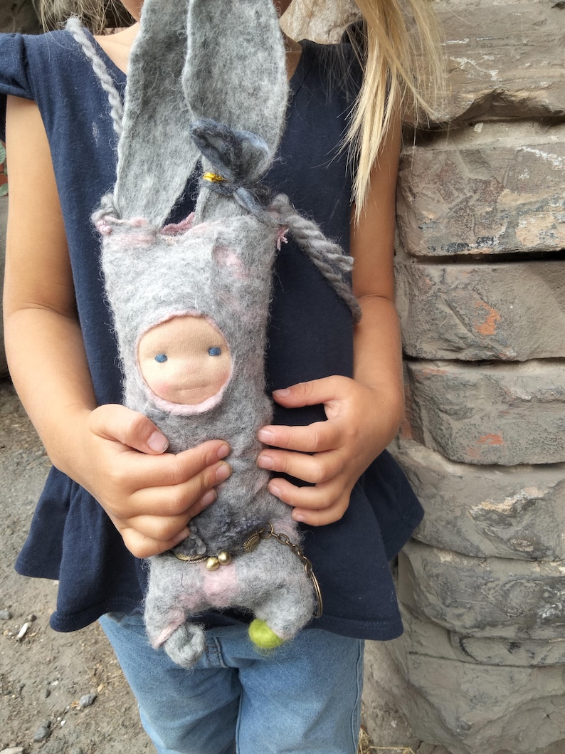 May include: A handmade gray felt bunny doll with long ears, a small face, and a rope strap. The doll has pink accents and a small green ball at the bottom. The doll is being held by a person wearing a navy blue top and blue jeans.