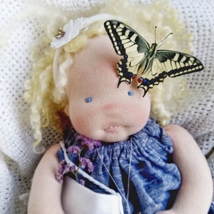 Puede incluir: Muñeca hecha a mano con cabello rubio rizado y ojos azules, vestida con un vestido azul. Una gran mariposa con marcas negras, amarillas y blancas descansa sobre la frente de la muñeca. La muñeca está acostada sobre una manta blanca texturizada.