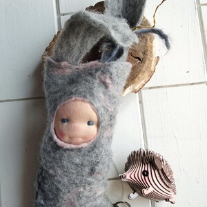May include: A handmade felt doll with a gray hooded outfit and a pink face. The doll has blue eyes and a decorative belt. A wooden hedgehog keychain is attached. The doll is approximately 8 inches tall.