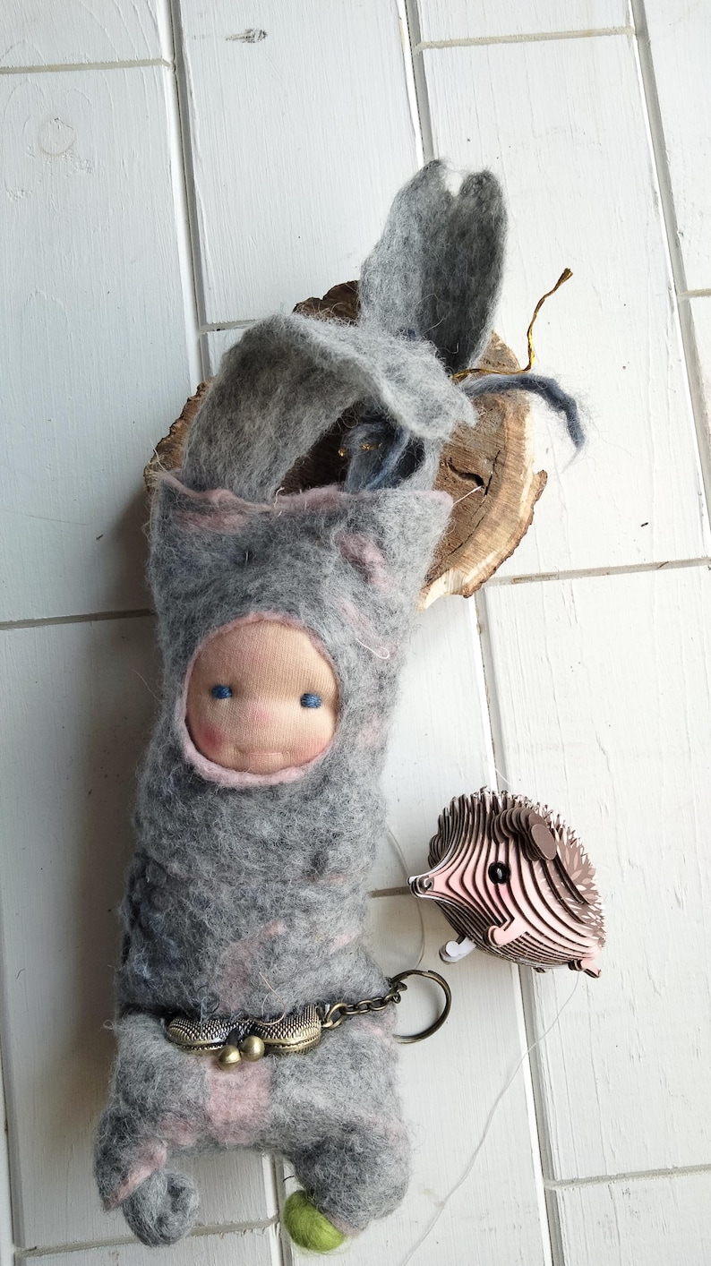 May include: A handmade felt doll with a gray hooded outfit and a pink face. The doll has blue eyes and a decorative belt. A wooden hedgehog keychain is attached. The doll is approximately 8 inches tall.
