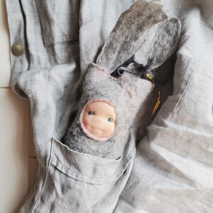 May include: A handmade gray felt doll with a pink-trimmed face and blue eyes peeks out of a gray pocket. The doll has long, floppy ears and is nestled in a gray linen garment. The background is a soft, textured gray fabric.