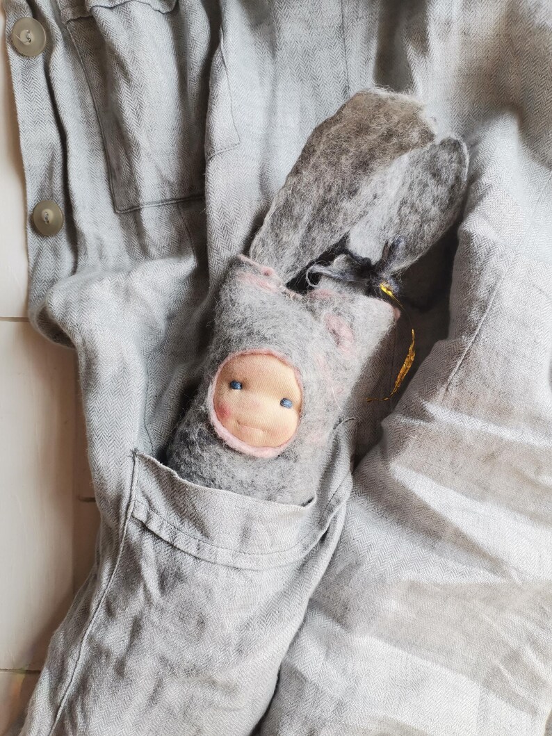 May include: A handmade gray felt doll with a pink-trimmed face and blue eyes peeks out of a gray pocket. The doll has long, floppy ears and is nestled in a gray linen garment. The background is a soft, textured gray fabric.