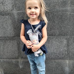 May include: A young child wearing a navy blue top, light blue jeans, and pink sandals. She is holding a gray doll with a white face and a gray hat. The child is smiling and looking upwards.