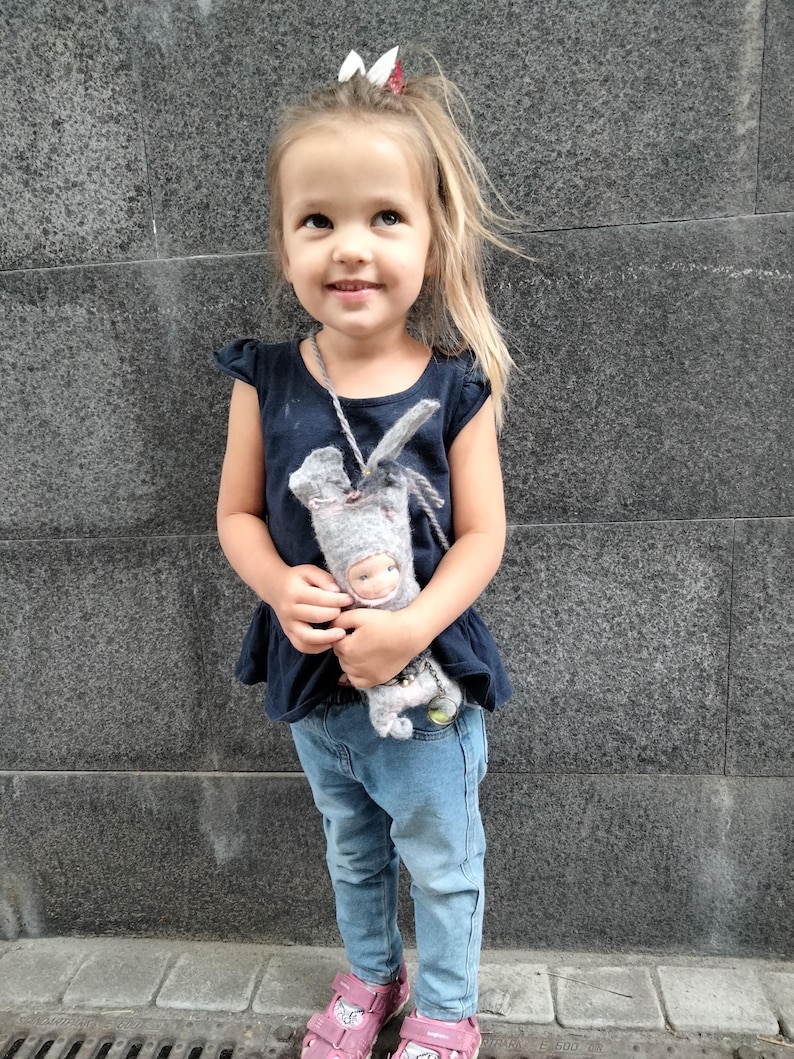 May include: A young child wearing a navy blue top, light blue jeans, and pink sandals. She is holding a gray doll with a white face and a gray hat. The child is smiling and looking upwards.