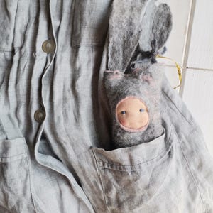 May include: A gray linen jacket with button closures and pocket details. A handmade gray felt doll with a pink face and blue eyes is nestled in the jacket pocket. The doll has long, floppy ears.