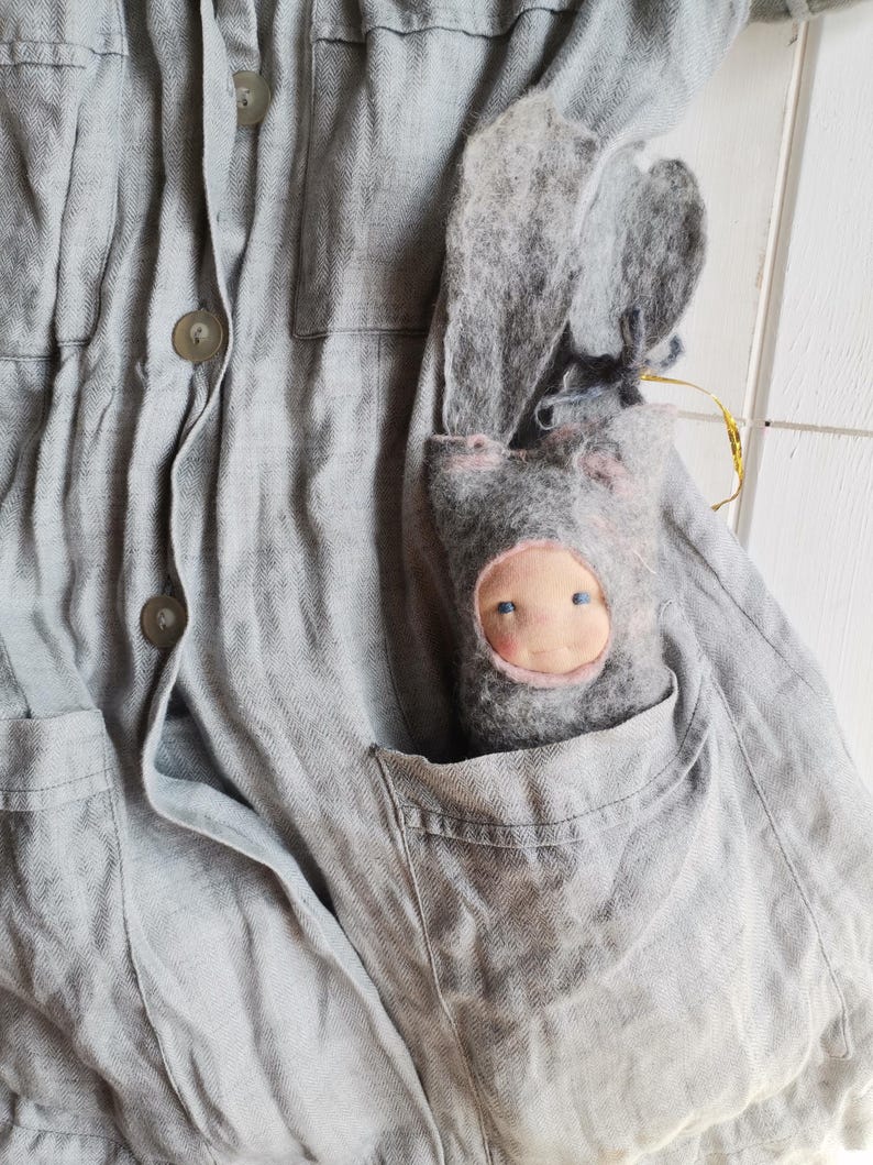 May include: A gray linen jacket with button closures and pocket details. A handmade gray felt doll with a pink face and blue eyes is nestled in the jacket pocket. The doll has long, floppy ears.