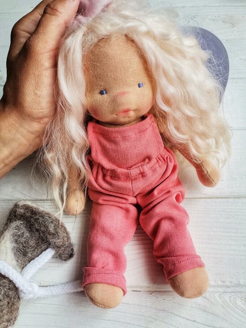 Pode incluir: Boneca de pano feita &agrave; m&atilde;o com cabelo loiro longo e ondulado e olhos bordados azuis. A boneca usa um macac&atilde;o rosa e tem p&eacute;s cor de pele. A boneca est&aacute; sendo segurada por uma m&atilde;o. Um pequeno animal de pel&uacute;cia cinza e branco est&aacute; no canto inferior esquerdo.