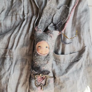 May include: A handmade felt doll in the shape of a bunny, with long ears and a pink string strap. The doll has a light blue face and is wearing a gray and pink outfit. A decorative metal clasp is attached to the doll.
