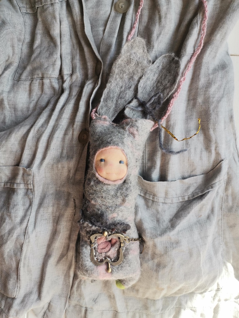 May include: A handmade felt doll in the shape of a bunny, with long ears and a pink string strap. The doll has a light blue face and is wearing a gray and pink outfit. A decorative metal clasp is attached to the doll.
