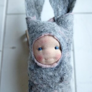 May include: A handmade doll with a gray felt bunny hood and a soft, pink-cheeked face. The doll has blue eyes and a gentle smile. The hood has long, floppy ears. A charming, whimsical toy.