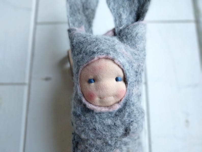 May include: A handmade doll with a gray felt bunny hood and a soft, pink-cheeked face. The doll has blue eyes and a gentle smile. The hood has long, floppy ears. A charming, whimsical toy.