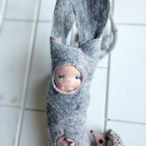May include: A handmade felt doll with a light blue face and blue eyes, wrapped in a gray felt bunny suit with long ears. A small felt hedgehog is next to the doll. The doll has a pink rose detail.