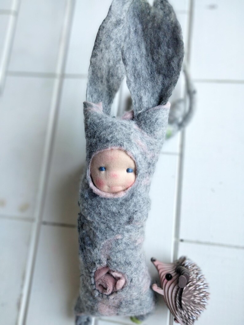 May include: A handmade felt doll with a light blue face and blue eyes, wrapped in a gray felt bunny suit with long ears. A small felt hedgehog is next to the doll. The doll has a pink rose detail.