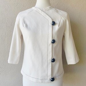 Puede incluir: Una chaqueta blanca de manga corta, abotonada, con cierre diagonal y botones negros.