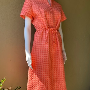 Op de afbeelding: Een koraalroze jurk met een V-hals en korte mouwen. De jurk heeft een strikceintuur in de taille en een geometrisch patroon.