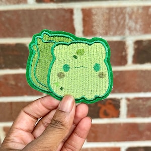 May include: Green embroidered iron-on patch of a cartoon character resembling a piece of bread with a smiling face.