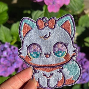 May include: A white embroidered patch of a cartoon cat with a pink bow, blue eyes, and a light blue tail. The cat is wearing a purple collar.