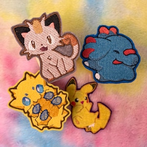 May include: Four embroidered patches featuring cartoon characters. One is a beige cat-like creature, one is a blue blob-like figure with red accents, one is a yellow spiky character with blue spots, and one is a yellow mouse-like character.