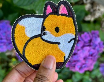 May include: A yellow and white embroidered fox patch with black outlines and pink ears. The patch is held in a hand against a background of purple flowers.