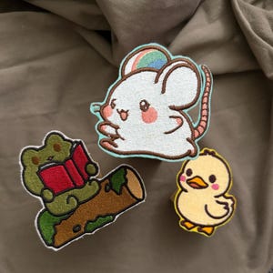 May include: Three embroidered patches featuring a white mouse with a rainbow on its head, a green frog reading a red book while sitting on a brown log, and a small yellow duckling with orange feet and a beak, all with distinct outlines.