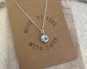 May include: A silver necklace featuring a small, round, textured pendant with the letter A stamped in the centre. The necklace is displayed on a brown card printed with the text Alice Stevens Metalsmith, which rests on a beige fabric pouch.